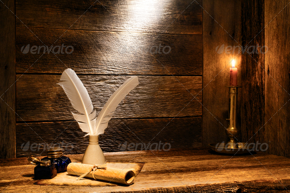 Antique parchment paper sheets roll tied by a string with old writing feather quills in a vintage ink well and ancient colonial blue glass inkwells on a distressed ancient wood desk lit by candlelight in an ancient historic home
