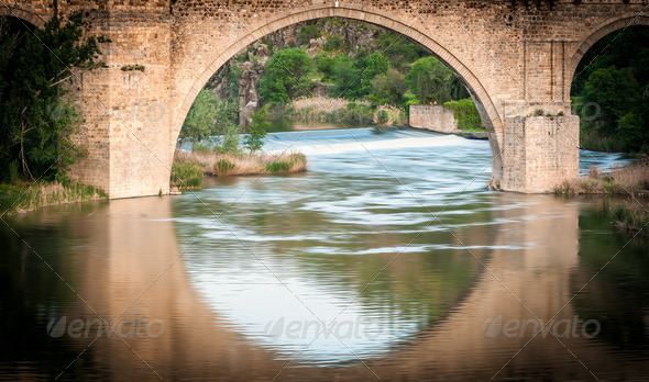 Famous bridge in Toledo, Spain. Arch and reflection in water form big circle. River running fast. Beautiful nature with green trees in background. Popular place for tourism in Europe.