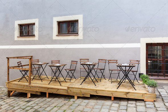 Empty restaurant or coffee shop terrace with wooden and metalic chairs and tables on the wooden platforim in front of the pink wall on the street covered with granite tiles