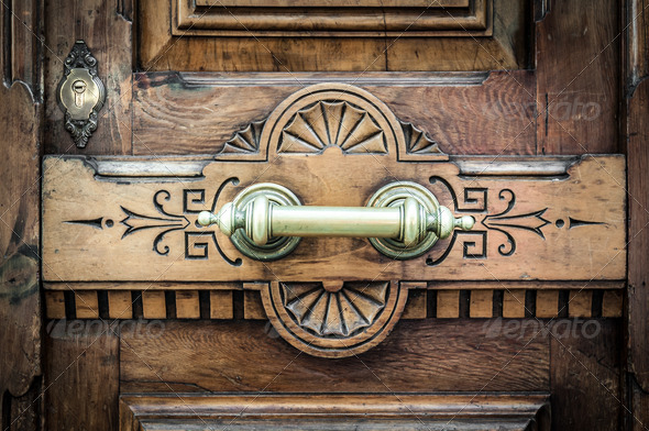 Detail of old wooden door with horizontal metal handle on board with carved pattern and keyhole over it. Dark brown solid front door in vintage and antique style. Exterior and facade.