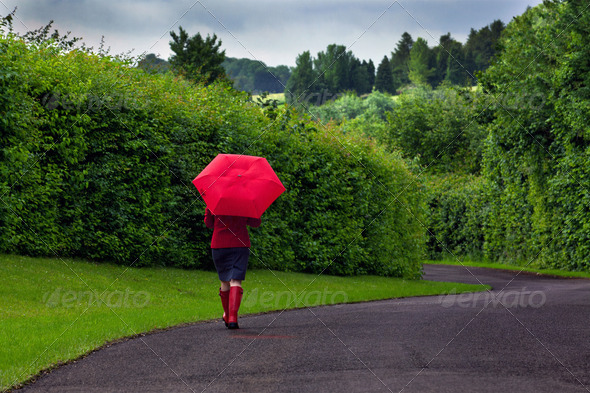 Photo of a woman walking down a road holding a red umbrella after a heavy downpour of rain on an overcast day.