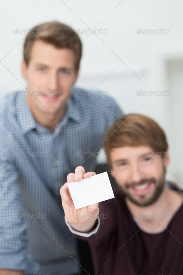 Businessman seated with a colleague holding out a blank business card fro your contact details with selective focus to the card