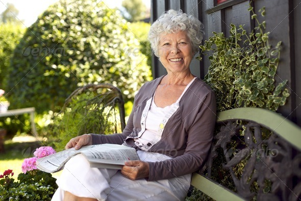 Happy senior woman sitting on bench in her backyard garden with a newspaper looking at camera smiling