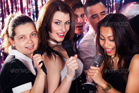 Portrait of beautiful women singing into microphones with male friends at karaoke party