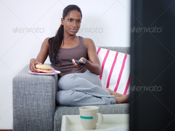 mid adult indian woman on sofa, changing tv channel with remote control and eating junk food. Front view, copy space