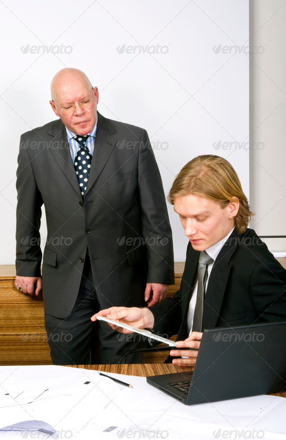 A senior manager looking down on a younger associate, working with a ruler and a laptop on several designs on the desk in front of him