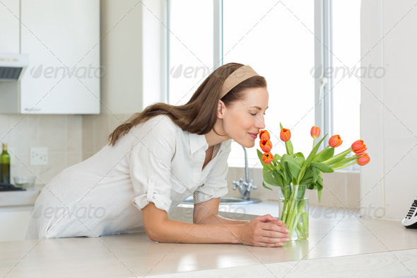 Smiling young woman smelling flowers in the kitchen at home (Misc) Photo Download