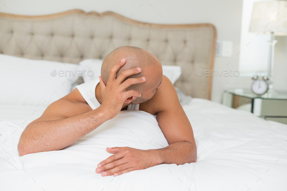 Sleepy young bald man looking at the alarm clock in bed at home