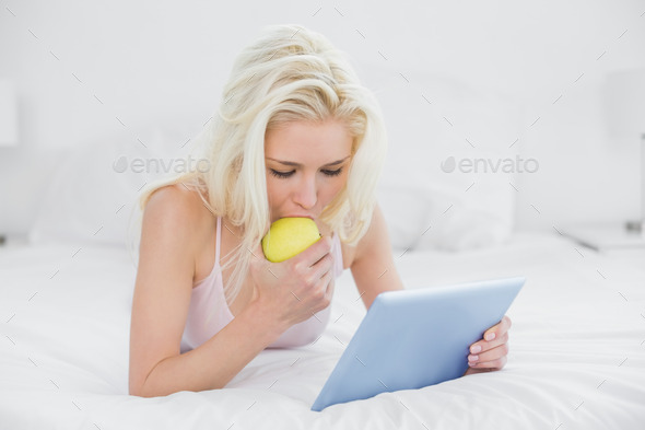 Casual young blond eating an apple while using tablet PC in bed at home
