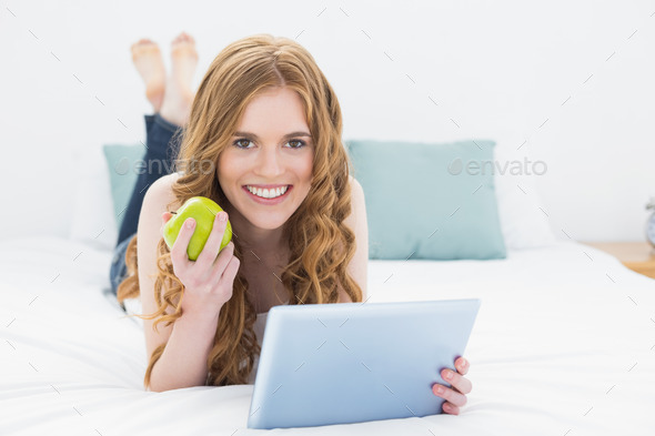 Portrait of a casual young blond using tablet PC while holding an apple in bed at home