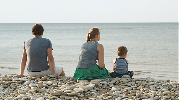Family Of Three Sitting On Pebble Beach By Water Family Of Three Sitting On Pebble Beach By Water