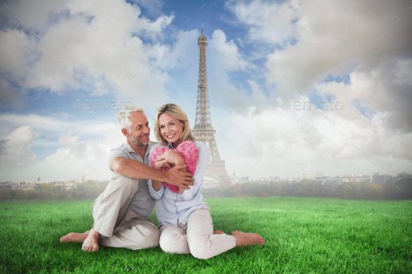 Happy couple sitting and holding heart pillow against eiffel tower (Misc) Photo Download