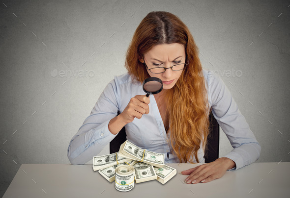 woman looking through magnifying glass on stack of dollar banknotes