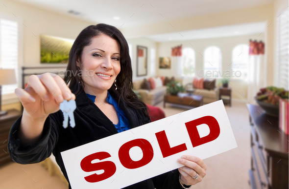 Happy Hispanic Woman with House Keys and Sold Sign in Beautiful Living Room.