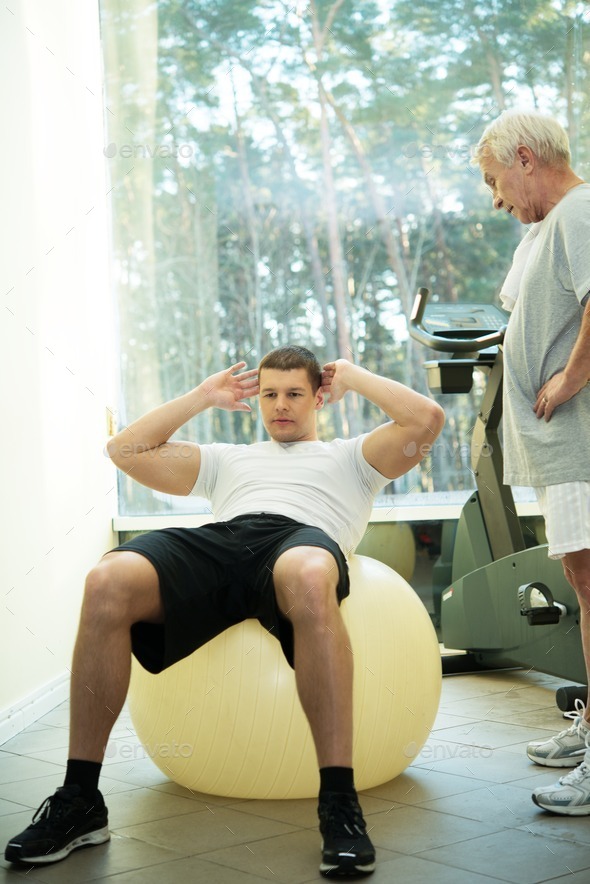 Personal trainer shows to a senior man how to do exercise on a fitness ball