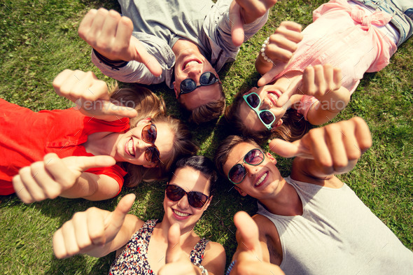 smiling friends showing thumbs up lying on grass