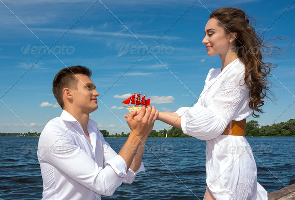 The guy is kneeling before the girl near the water